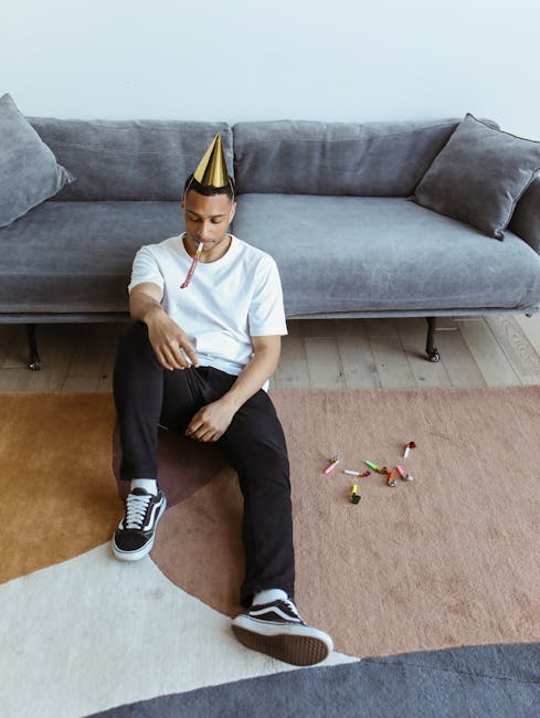 A young person sitting on a multicolored carpet in a living room at a party, wearing a gold party hat and holding a party blower in their mouth, with discarded party poppers and confetti scattered around. The room features a gray fabric sofa with matching cushions against a plain white wall, and light wooden flooring. The lighting is natural and soft, highlighting the clean and well-maintained appearance of the carpeted surface. The scene suggests post-party cleaning, and the image reflects the importance of surface cleaning and stain removal in maintaining hygiene and freshness in a domestic setting, as offered by Carpet Cleaning Kennington.