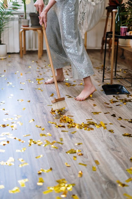 A young person sitting on a multicolored carpet in a living room at a party, wearing a gold party hat and holding a party blower in their mouth, with discarded party poppers and confetti scattered around. The room features a gray fabric sofa with matching cushions against a plain white wall, and light wooden flooring. The lighting is natural and soft, highlighting the clean and well-maintained appearance of the carpeted surface. The scene suggests post-party cleaning, and the image reflects the importance of surface cleaning and stain removal in maintaining hygiene and freshness in a domestic setting, as offered by Carpet Cleaning Kennington.