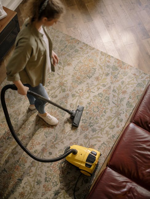 A person using a yellow vacuum cleaner with a long hose attachment to clean a patterned area rug in a living room. The room features light wooden flooring and a red leather sofa positioned along the edge of the rug. The individual is dressed in casual clothing, including a beige jacket and light-colored shoes, and is focused on surface cleaning. The scene is well-lit, emphasizing the cleanliness and hygiene of the carpet and surrounding surfaces, which reflects professional deep cleaning and sanitisation services provided by Carpet Cleaning Kennington as part of their domestic cleaning offering in the SE11 area.