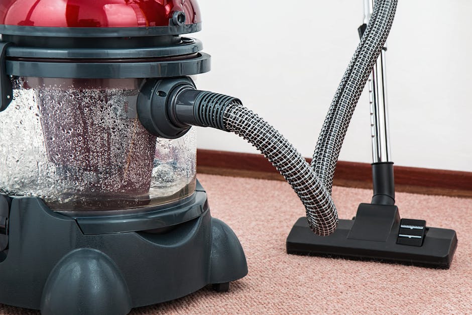 Close-up of a portable vacuum cleaner with a clear dust container, filled with debris and bubbles, connected to a patterned hose and a black cleaning head on a plush pink carpet in a well-lit room. The vacuum is positioned near a white wall with a brown baseboard. Visible parts include the red lid, grey body, and black base, highlighting surface cleaning and maintenance focused on deep cleaning and sanitisation of domestic carpets. The setting suggests a professional cleaning environment with a focus on hygiene and surface cleanliness, as managed by Carpet Cleaning Kennington.