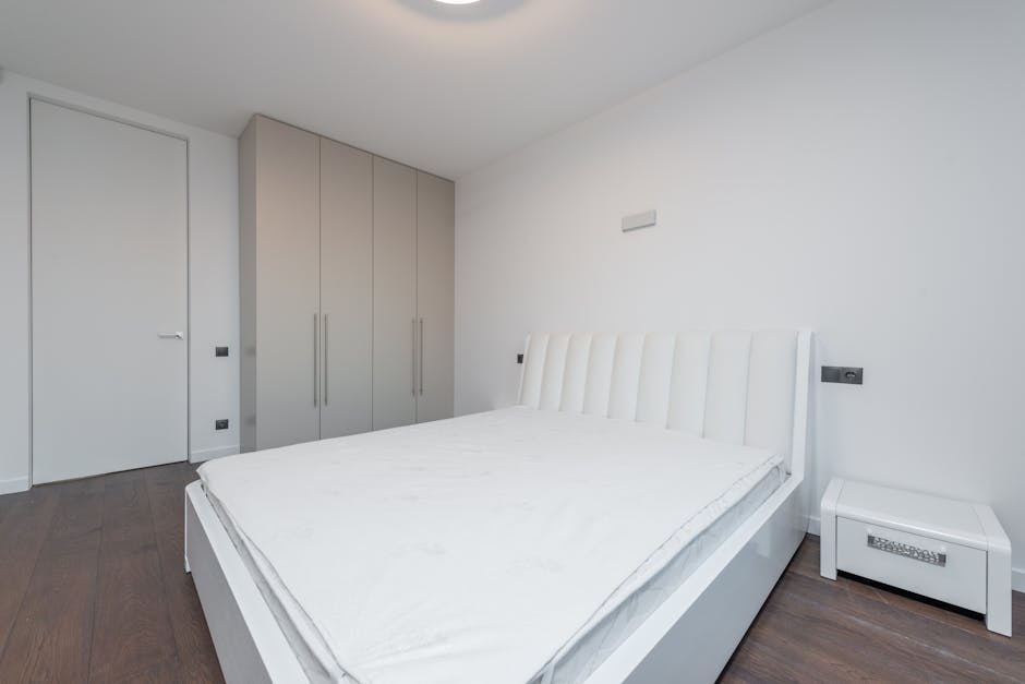A person is making a bed with white, neatly pressed sheets and a pillow on a wooden bed frame in a bedroom. The individual is in the process of adjusting or arranging the bedding, with their hand plac