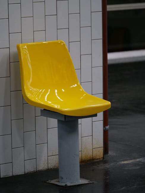 A bright yellow plastic seat mounted on a metal pedestal is situated against a white tiled wall inside a station. The tiles are rectangular and arranged in a vertical pattern with a slight sheen, indicating they are clean and well-maintained. The seat appears glossy and free of dirt or marks, suggesting recent surface cleaning. The surrounding area shows a dark, possibly tiled or concrete floor, with a small rusty stain near the base of the pedestal. In the background, a glass panel and a part of the station interior are faintly visible, illuminated by subdued lighting. This setting is within an indoor station environment, matching the theme of surface cleaning and maintenance services offered by Carpet Cleaning Kennington, as referenced in the page titled 'Oval station upholstery cleaning for Kennington commuters'.