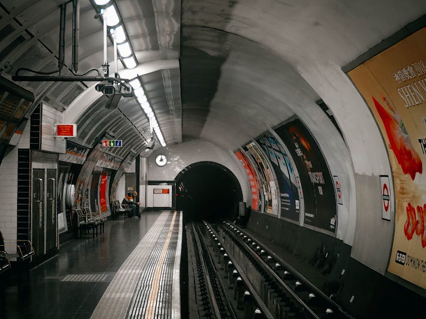 Indoor view of the upward escalator and train tracks at Oval station, part of the London Underground, featuring curved white tiled walls, illuminated ceiling lighting, and a large advertising poster on the right side. The platform area appears clean and well-maintained, with a seating bench, signage for exits and facilities, and a few commuters visible in the distance, all within a modern, underground transit setting. Carpet Cleaning Kennington offers surface cleaning and hygiene maintenance for similar commercial environments.