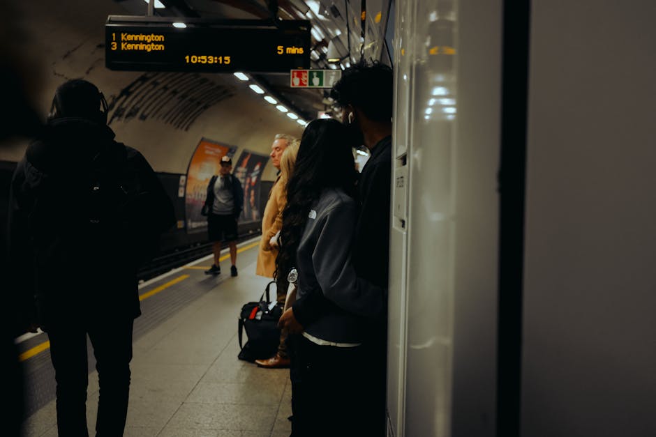 A bright yellow plastic seat mounted on a metal pedestal is situated against a white tiled wall inside a station. The tiles are rectangular and arranged in a vertical pattern with a slight sheen, indicating they are clean and well-maintained. The seat appears glossy and free of dirt or marks, suggesting recent surface cleaning. The surrounding area shows a dark, possibly tiled or concrete floor, with a small rusty stain near the base of the pedestal. In the background, a glass panel and a part of the station interior are faintly visible, illuminated by subdued lighting. This setting is within an indoor station environment, matching the theme of surface cleaning and maintenance services offered by Carpet Cleaning Kennington, as referenced in the page titled 'Oval station upholstery cleaning for Kennington commuters'.