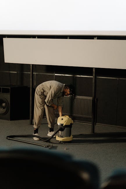 A person in a light-colored jumpsuit is leaning over and operating a yellow carpet cleaning machine on a dark grey carpeted floor. The room appears to be a conference or presentation space with black wall panels, a large white projection screen, and an air vent below it. There are visible audio equipment, including a speaker, positioned near the wall. The area is well-lit, and the cleanliness of the carpet suggests deep cleaning. The scene relates to surface cleaning and sanitation services offered by Carpet Cleaning Kennington, demonstrating professional deep cleaning processes for commercial spaces.