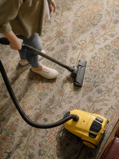 A person wearing casual clothing, including beige shoes and dark pants, is using a vacuum cleaner with a black hose attachment to clean a traditional patterned carpet in a room. The carpet features a floral design with muted tones of beige, green, and coral. The yellow vacuum cleaner, positioned on the floor, has a black power cord connected to it. The room has natural lighting, illuminating the carpet's detailed design, and the cleaning process is part of a deep surface cleaning procedure. Carpet Cleaning Kennington specializes in domestic cleaning and surface sanitation, ensuring carpets are thoroughly cleaned and hygienic.