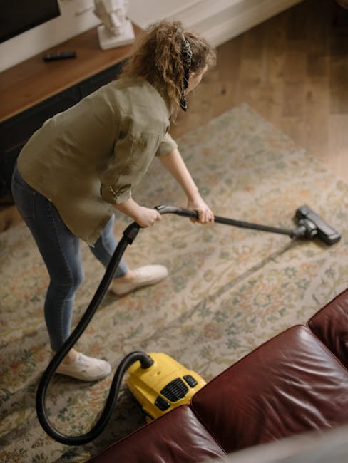 A person in a light-colored jumpsuit is leaning over and operating a yellow carpet cleaning machine on a dark grey carpeted floor. The room appears to be a conference or presentation space with black wall panels, a large white projection screen, and an air vent below it. There are visible audio equipment, including a speaker, positioned near the wall. The area is well-lit, and the cleanliness of the carpet suggests deep cleaning. The scene relates to surface cleaning and sanitation services offered by Carpet Cleaning Kennington, demonstrating professional deep cleaning processes for commercial spaces.