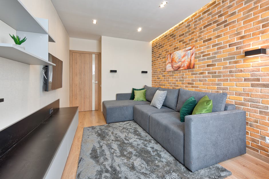 A modern living room with a grey fabric sectional sofa adorned with green and grey cushions, situated on a textured grey rug. The room features a prominent exposed brick wall decorated with a colorful abstract painting and two black wall-mounted lights. To the left, white floating shelves hold a potted plant and electronic devices, with a black surface below. The walls are painted in neutral tones, and a wooden door with frosted glass is visible in the background. The space is well-lit with ceiling spotlights, and the surfaces appear clean and well-maintained, reflecting professional domestic cleaning standards by Carpet Cleaning Kennington.