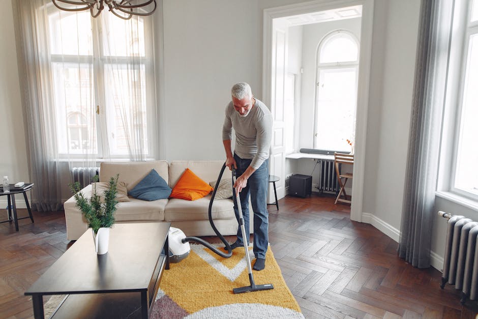 A middle-aged man with grey hair and beard, dressed in a light grey sweater and dark trousers, is performing surface cleaning in a spacious, well-lit living room. He is using a vacuum cleaner with a hose attachment to clean a yellow area rug with a modern geometric pattern. The room features large windows with sheer white curtains allowing natural daylight to fill the space. The wooden floor has a polished, dark finish, contrasting with the white walls and trim. A white sofa with colorful cushions (blue, orange) is positioned against the wall, with a small side table holding a glass of water nearby. A white coffee table with a small potted plant sits in the foreground, while a compact black heater or speaker, a wooden desk, and a chair are visible in the background. The room appears meticulously maintained and clean, highlighting effective domestic cleaning practice, with the attention to hygiene and surface sanitisation characteristic of professional services offered by Carpet Cleaning Kennington.